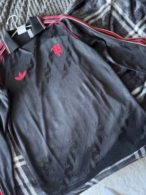 Manchester United tröja - Säljer en svart långärmad skjorta från Adidas med diskret mönster av Adidas-loggor i tyget. Skjortan har röda detaljer på axlar och krage samt broderade loggor på bröstet. Perfekt för dig som gillar sportig stil och snygga detaljer.