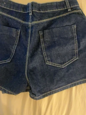 Mörkblå denimshorts med fickor - Snygga mörkblå denimshorts med stora fram- och bakfickor samt kontrastsömmar. Klassisk jeanslook med bälteshällor och knappstängning framtill. Perfekta för sommarens alla äventyr.