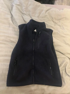Mörkblå fleeceväst  - Mysig mörkblå fleeceväst med hög krage och tre dragkedjeförsedda fickor framtill. Västen har en enkel och stilren design, perfekt att slänga på sig över en hoodie eller tröja. Materialet är mjukt och värmande fleece.