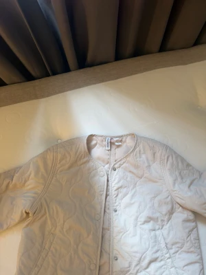 Beige quiltad cardiganjacka - Stilren beige quiltad cardiganjacka med rund halsringning och tryckknappar framtill. Jackan har ett vågigt mönster i quiltningen och två fickor framtill. Perfekt för lager på lager och enkel att matcha med olika outfits.