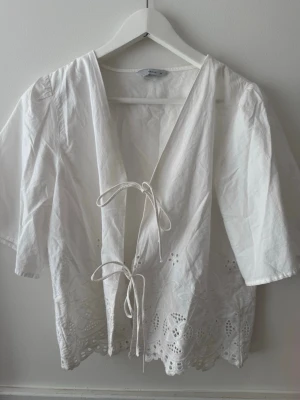 Vit blus med knyt och broderi - Söt vit blus med v-ringning, knyt framtill och broderade detaljer längs nederkanten. Blusen har korta ärmar och ett luftigt material som passar perfekt till jeans eller kjol. Spetsdetaljer ger en romantisk touch.