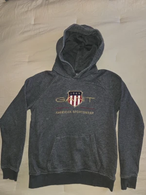 Mörkgrå hoodie från GANT - Snygg mörkgrå hoodie från GANT med broderad logga och texten 'American Sportswear' på bröstet. Klassisk känguruficka framtill och mjukt material som känns skönt mot huden. Perfekt för en avslappnad och sportig stil.