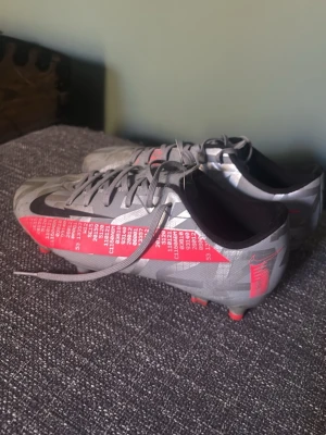 Nike grå/röda Mercurial fotbollsskor - Snygga Nike Mercurial fotbollsskor i grått med röda och svarta detaljer. Skorna har snörning, låg profil och mönstrad ovandel med text och grafik på sidan. Perfekta för snabba spelare som vill sticka ut på planen.