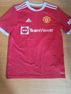 Manchester United matchtröja Elanga - Röd Manchester United matchtröja från Adidas med vita detaljer, klubbmärke och TeamViewer-logga på bröstet. Nummer 36 och namnet Elanga på ryggen. Tillverkad i lätt och ventilerande material, perfekt för fotbollsträning eller match.