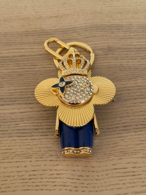 Vuitton Guldig nyckelring med strass och krona - Unik nyckelring i form av en figur med guldtonad metall, dekorerad med gnistrande strass och en stor krona. Figuren har blå ben och detaljer i svart och vitt. Perfekt accessoar för att piffa upp väskan eller nyckelknippan.