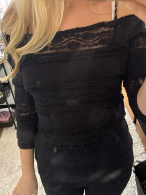 Svart offshoulder topp i spets - Snygg svart offshoulder topp med spetsdetaljer och trekvartsärm. Toppen har en figurnära passform och är perfekt för dig som gillar en trendig och stilren look. Spetsen ger en extra touch och gör toppen unik.