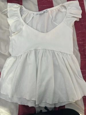 Vit blus med volangärmar - Supersöt vit blus med volangärmar och v-ringning. Toppen har en lös passform och är tillverkad i ett lätt, glansigt material som ger en fräsch känsla. Perfekt att styla med jeans eller kjol för en trendig look.