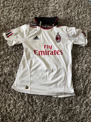Vit AC Milan matchtröja Adidas - Snygg vit AC Milan matchtröja från Adidas med klubbmärke och Fly Emirates-tryck på bröstet. Tröjan har svarta och röda detaljer vid kragen och axlarna samt korta ärmar. Tillverkad i funktionsmaterial som passar perfekt till fotbollsträning eller match.