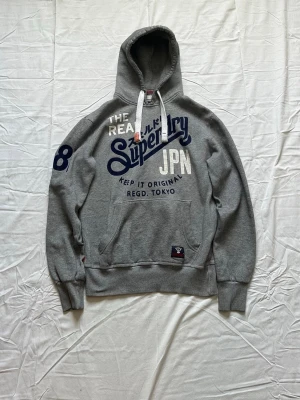 Grå hoodie från Superdry med tryck - Snygg grå hoodie från Superdry med stort blått och vitt tryck på bröstet och ärmen. Klassisk känguruficka framtill, vita dragsnören i huvan och ribbade muddar. Perfekt för en avslappnad streetstyle-look.