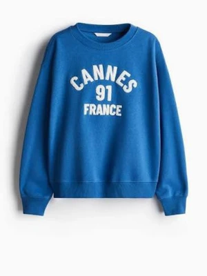 Blå sweatshirt med Cannes-tryck - Cool blå sweatshirt med texten 'CANNES 91 FRANCE' i vitt framtill. Tröjan har rund halsringning, ribbade muddar vid ärmslut och nederkant samt en avslappnad passform. Perfekt för en casual och sportig look.