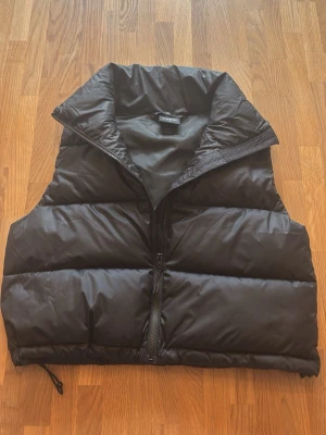 Svart puffad dunväst med hög krage - Snygg svart dunväst med puffad design och hög krage. Västen har dragkedja framtill och justerbar nederkant med snöre. Perfekt för lager på lager och ger en cool streetwear-look.