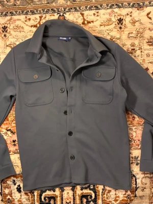 Mörkgrå overshirt med fickor - Stilren mörkgrå overshirt med klassisk krage och fyra knappar framtill. Två stora bröstfickor med lock och knapp. Tillverkad i ett mjukt, stretchigt material som ger en avslappnad look. Perfekt att bära öppen över en t-shirt.