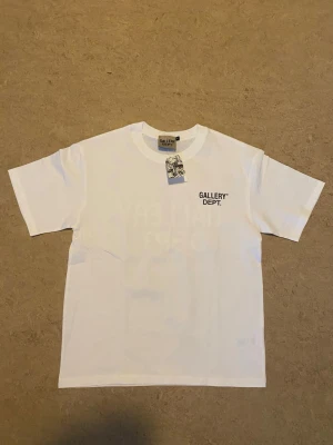 Vit Gallery Dept Tee - Det här är en klassisk Gallery Dept T-shirt som är helt ny med tags och påse. Allt följer med vid köp! Den är i storlek M och är true to size. Denna t-shirten är perfekt om du är ute efter streetwear stil eller bara för en vardags look, eller om man vill kan man ha den på specifika tillfällen. 