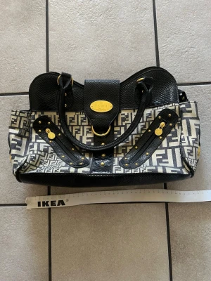 Fendi Selleria svart och beige handväska - Snygg handväska från Fendi Selleria i svart och beige med klassiskt FF-mönster. Väskan har detaljer i svart läder med guldfärgade metalldelar och dubbla handtag. Perfekt för dig som vill ha en statementväska med lyxig känsla och unik design.