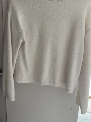 Vit fluffig stickad tröja - Mysig vit stickad tröja med lång ärm och rund hals. Tröjan har en fluffig struktur och ribbstickade muddar vid ärmslut och nederkant. Perfekt för kyliga dagar och enkel att matcha med olika outfits.