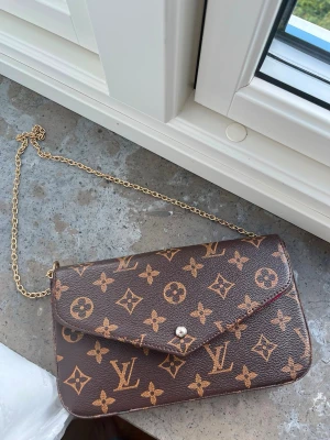 Väska  - Snygg brun kuvertväska med klassiskt monogrammönster i guld och beige. Väskan har guldfärgad kedja och röd insida. Tillverkad i tåligt lädermaterial och stängs med tryckknapp. Perfekt accessoar för att lyfta din outfit.
