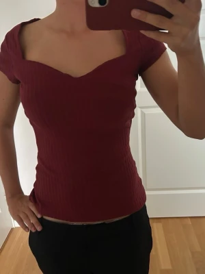 Mörkröd ribbad topp med v-ringning - Snygg mörkröd ribbad topp med korta ärmar och unik v-ringad hals. Toppen har en figurnära passform och är tillverkad i ett stretchigt material som sitter bekvämt. Perfekt för dig som vill ha en enkel men trendig look. Toppen är från Anna field!😊