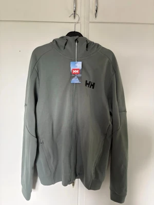Grå hoodie med dragkedja från Helly Hansen - Snygg grå hoodie från Helly Hansen med hel dragkedja framtill och svart HH-logga på bröstet. Hoodien har huva med dragsko och två sidofickor. Tillverkad i mjukt material som passar perfekt för en avslappnad stil. Helt ny 