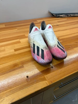 Adidas vita och rosa fotbollsskor - Säljer ett par Adidas fotbollsskor i vitt med rosa och svarta detaljer. Skorna har en strömlinjeformad design utan snörning och tydliga dobbar för bra grepp på planen. Perfekta för dig som vill ha stilrena och snabba skor på matchen.