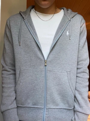 Grå hoodie från Polo Ralph Lauren - Snygg grå zip-hoodie från Polo Ralph Lauren med huva och dragsko. Klassisk logga broderad på bröstet och två stora fickor framtill. Tillverkad i mjukt bomullsmaterial, perfekt för en chill och stilren look.