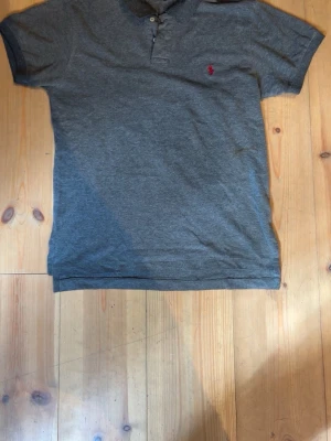 Grå piké från Polo Ralph Lauren - Säljer en klassisk grå pikétröja från Polo Ralph Lauren i bomull. Tröjan har korta ärmar, krage med knappar och den ikoniska röda loggan broderad på bröstet. Passformen är custom fit vilket ger en snygg siluett.