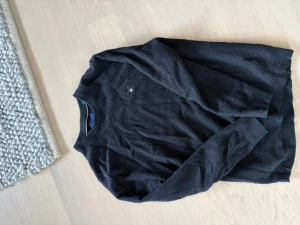 Mörkgrå stickad tröja från GANT - Snygg mörkgrå stickad tröja från GANT med rund halsringning och diskret broderad logga på bröstet.  Den är i merinoull. Tröjan har långa ärmar och är tillverkad i mjukt material som passar perfekt till lager på lager. Klassisk och stilren design. Det står XL på tröjan men på grund av vårdslös tvättning har tröjan fått en ny passform som liknar small (S).