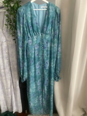 Blommig blågrön klänning från Adoore - Säljer en lång, blågrön klänning från Adoore med blommigt mönster i lila och blått. Klänningen har djup v-ringning, långa puffärmar och knappar framtill. Materialet är glansigt och ger ett lyxigt intryck. Perfekt för dig som vill sticka ut med en romantisk look.