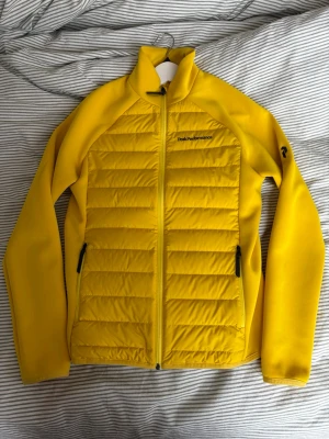 Gul pufferjacka från Peak Performance - Snygg gul pufferjacka från Peak Performance med quiltad framsida och släta ärmar. Jackan har hel dragkedja, två fickor med dragkedja och logga på bröstet samt ärmen. Perfekt för kalla vinterdagar och riktigt skön passform. Smal modell, känns som XS.