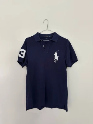Mörkblå pikétröja från Polo Ralph Lauren - Säljer en Ralph Lauren Big Pony pikétröja i modellen Custom Slim Fit Mesh. Klassisk och stilren piké i hög kvalitet med ikonisk Big Pony-logga. Använd endast cirka två gånger och är i närmast nyskick. Nypris ca 2 295 kr. Perfekt till både vardag och mer uppklädda tillfällen. Skickas eller hämtas enligt överenskommelse.