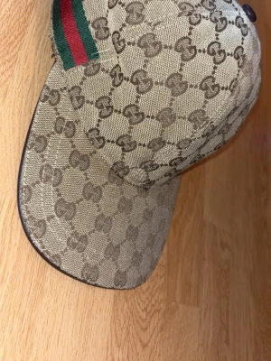 Beige Gucci keps (box finns) - Snygg beige keps från Gucci med klassiskt GG-monogram över hela kepsen och en grön-röd rand på sidan. Kepsen är tillverkad i canvasmaterial och har svart insida med etikett. Knappt använd så mycket bra skick!