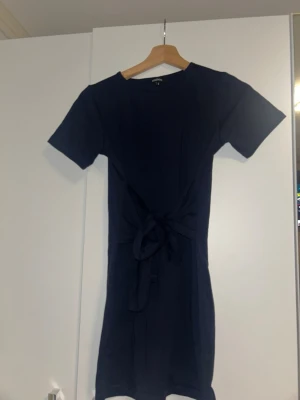 Mörkblå kort klänning från Boohoo - En enkel och stilren mörkblå klänning från Boohoo med korta ärmar och knytband i midjan. Klänningen har en rak passform och är tillverkad i ett mjukt material som känns bekvämt mot huden. Perfekt för dig som gillar minimalistisk stil.