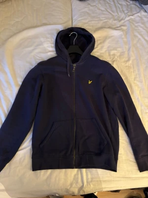 Marinblå hoodie från Lyle & Scott - Snygg marinblå hoodie från Lyle & Scott med dragkedja framtill och klassisk gul logga på bröstet. Tillverkad i ekologisk bomull och har två fickor framtill. Perfekt för en chill och stilren look. 