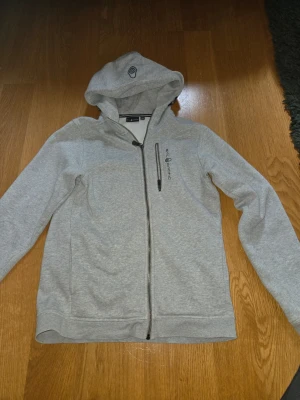 Grå hoodie från Sail Racing - Snygg grå hoodie från Sail Racing med dragkedja framtill och en bröstficka med zip. Hoodien har logga på bröstet och på huvan, samt mjukt material som känns skönt mot huden. Perfekt för chill dagar eller när du vill ha en clean look.