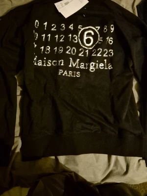 Maison Margiela svart hoodie - Säljer en svart hoodie från Maison Margiela med det ikoniska numermotivet och loggan tryckt i vitt på framsidan. Hoodien har en klassisk passform och är tillverkad i mjukt bomullsmaterial. Perfekt för dig som gillar streetwear och vill sticka ut.