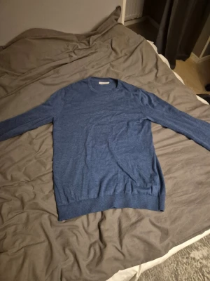 Blå tunn stickad tröja - Snygg blå tunnstickad tröja med rund halsringning och långa ärmar. Tröjan har ribbade muddar vid ärmslut och nederkant, vilket ger en clean och enkel look. Perfekt att matcha med jeans eller shorts för en avslappnad stil.