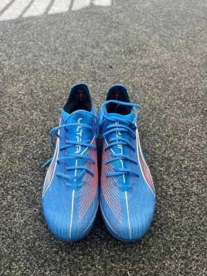  Puma Ultra fotbollsskor - Säljer ett par blåa Puma Ultra fotbollsskor med vita och röda detaljer. Skorna har snörning, låg profil och en lätt ovandel i syntetmaterial för maxad speed på planen. Perfekta för dig som vill sticka ut och få grym touch på bollen. Påse finns, pris kan förhandlas