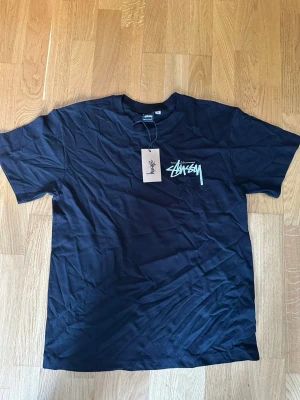 Svart Stüssy t-shirt med blå logga - Säljer en svart t-shirt från Stüssy med ikonisk blå logga tryckt på bröstet och stort på ryggen. Klassisk passform och rund hals. Perfekt för dig som gillar streetwear och vill ha något stilrent men ändå med edge.