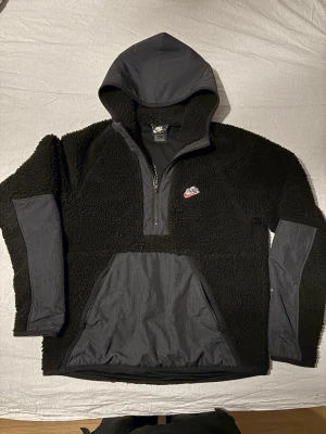 Nike fleece anorak  - Mysig svart fleecehoodie från Nike med halv dragkedja (anorak)  stor ficka framtill och huva i nylonmaterial. Tröjan har detaljer i nylon på ärmar och ficka samt en liten Nike-logga på bröstet. Perfekt för kalla dagar och streetwear-stil. Skriv så löser vi något bra. 