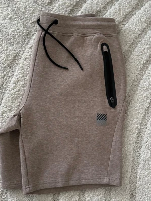 Beige shorts från Superdry med snörning - Säljer ett par beige shorts från Superdry med svart snörning i midjan och en dragkedjeförsedd ficka på sidan. Sköna och stilrena, perfekta för sommaren. Liten logga framtill och mjukt material.