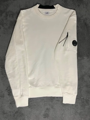 C.P. Company – White Lens Sweatshirt (M) -  Stylish and minimalist C.P. Company sweatshirt in clean white. Size M, length 66 cm, pit-to-pit 56 cm – fits perfectly for a modern, relaxed look. The sleeve features the iconic lens detail, instantly recognizable as C.P. Company’s signature.  ✅ Condition: Brand new – no wear, no stains 💯 Authenticity: 100% genuine, can be verified easily at certilogo.com   📦 Shipping: Dispatched immediately after purchase – fast and hassle-free!
