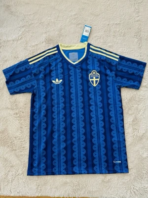 Blå Sverige fotbollströja Adidas - Säljer en blå fotbollströja från Adidas med Sveriges landslagsemblem på bröstet. Tröjan har ett mönster i olika blå nyanser, gula detaljer på axlarna och runt kragen samt korta ärmar. Perfekt för match eller träning!