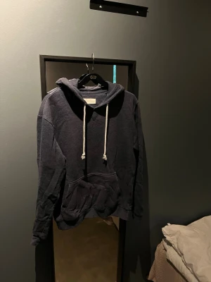 Mörkblå hoodie med vita snören - En stilren mörkblå hoodie med stora fickor framtill och vita dragsnören i huvan. Hoodien har en klassisk passform och är tillverkad i mjukt material som känns skönt mot huden. Perfekt för en avslappnad look och enkel att matcha med jeans eller joggers.
