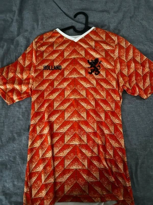 Holland retro fotbollströja orange från Amsterdam - Säljer en ikonisk orange Holland retro fotbollströja med geometriskt mönster, svart lejonemblem och texten 'HOLLAND' på bröstet. På ryggen står det 'GULLIT' och nummer 10 i svart. Tröjan har vit krage och är kortärmad, perfekt för dig som älskar klassiska fotbollströjor.