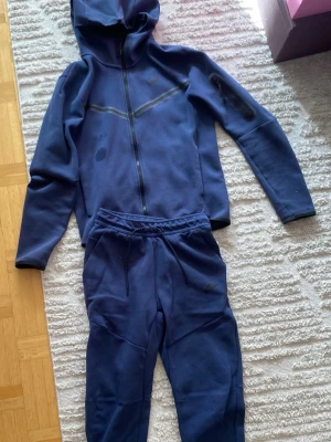 Mörkblå Nike loungewear-set - Snyggt mörkblått loungewear-set från Nike med mjuk hoodie och matchande byxor. Hoodien har dragkedja och byxorna har dragsko i midjan samt Nike-logga på benet. Perfekt för chill dagar hemma eller när du vill vara bekväm.