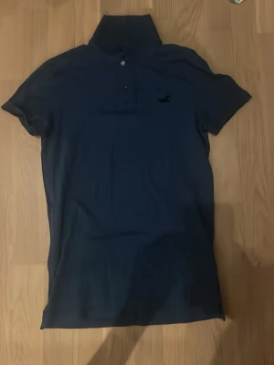 Blå pikétopp från Hollister - Mörkblå pikétopp från Hollister med klassisk krage och två knappar framtill. Toppen har korta ärmar och är tillverkad i ett stretchigt bomullsmaterial. Liten Hollister-logga broderad på bröstet.