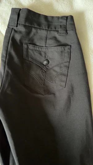 Svarta kostymbyxor från Gina Tricot XS - Säljer ett par svarta kostymbyxor från Gina Tricot i storlek XS. Jeansen har en bakficka med dekorativ knapp och söm, samt hällor för skärp. Materialet känns mjukt och stretchigt, perfekt för en snygg och bekväm look.