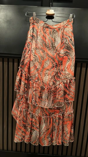 Mönstrad volangkjol i orange och beige - Säljer en långkjol med volanger och ett färgstarkt mönster i orange, beige och svart. Kjolen har flera lager volanger och ett flowy fall som ger en cool och trendig look. Perfekt för dig som vill sticka ut lite extra.