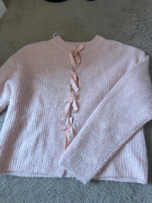 Ljusrosa stickad tröja med rosetter - Supersöt ljusrosa stickad tröja med fyra dekorativa rosetter framtill. Tröjan har en rak passform, lång ärm och ribbad struktur. Perfekt för dig som gillar gulliga detaljer och mjuka material. I verkligheten är tröjan mer rosa men det syns inte på bilderna