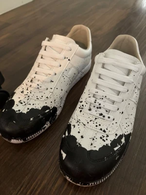 Maison marigela skor - Unika vita sneakers med svart färgstänksdesign över tå och sidor. Klassisk låg modell med vita snören och sula i gummi. Skorna har en modern och edgy look som sticker ut. Perfekta för dig som gillar streetstyle och vill ha något annorlunda.