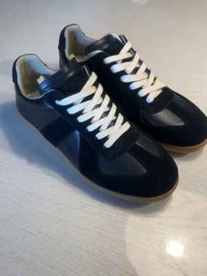 Maison margiela sneakers - Stilrena svarta sneakers med vita snören och beige sula. Skorna har en mix av mocka och slätt läder, samt klassisk låg profil. Perfekta för dig som gillar en clean och tidlös look.